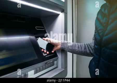 Mann`s Hand mit einem Smartphone mit einem Geldautomaten an der Straße, um etwas Geld abzuheben Stockfoto